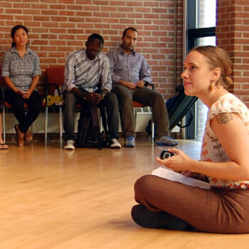 A woman sitting cross-legged on hardwood floor. There are four people sitting on chairs against a brick wall behind her.