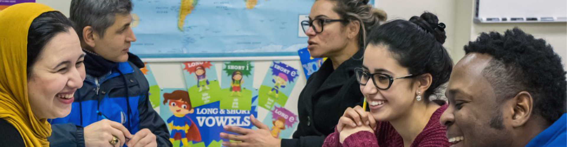 A group of immigrant students in a classroom smiling, talking and laughing with one another.