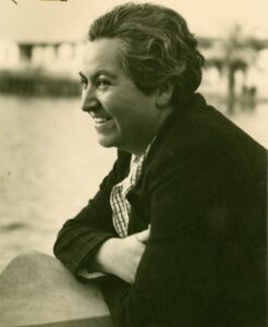 Gabriela Mistral leaning on a wall and staring out into some water. She has short gray hair and is wearing a thick coat.