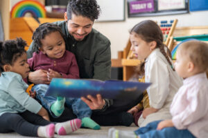 A male daycare educator of Hispanic decent sits on the floor with a child on his lap and other toddlers scattered around him, as he reads them a story. They are all focused on the book.