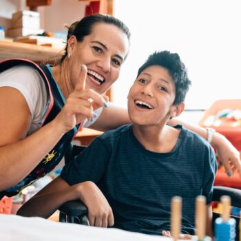 A woman smiling with a young boy with a disability, who is also smiling. They're in a classroom.