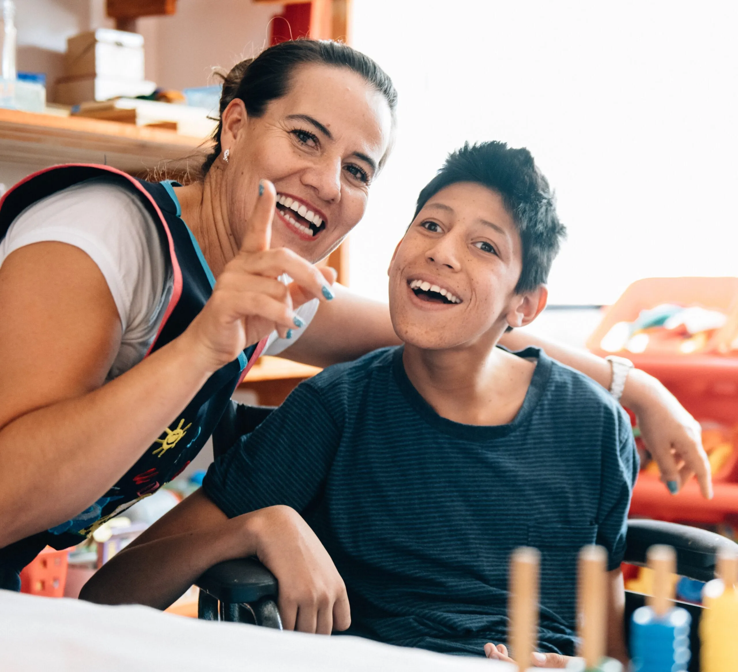 A woman smiling with a young boy with a disability, who is also smiling. They're in a classroom.