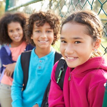 Three kids smiling at the camera. They're leaning on a wire fence and wearing backpacks.