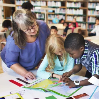 A woman leaning over two kids who are looking at a workbook on a desk.