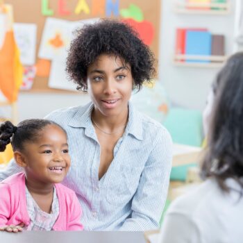 A woman and her young daughter, who is smiling, in a classroom talking with a teacher.
