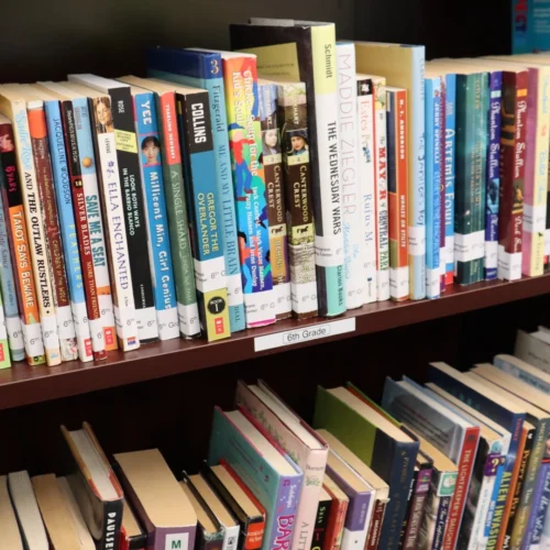Dozens of books on a library shelf that's labeled for sixth grade. They all have Immigrant Learning Center labels on the spines.