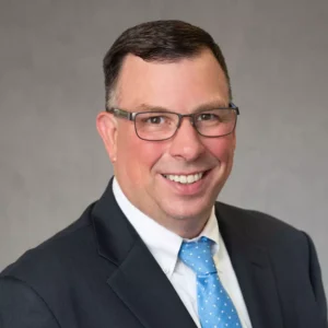 Vince Rivers, a man with dark hair and glasses wearing a dark suit jacket and light blue tie.