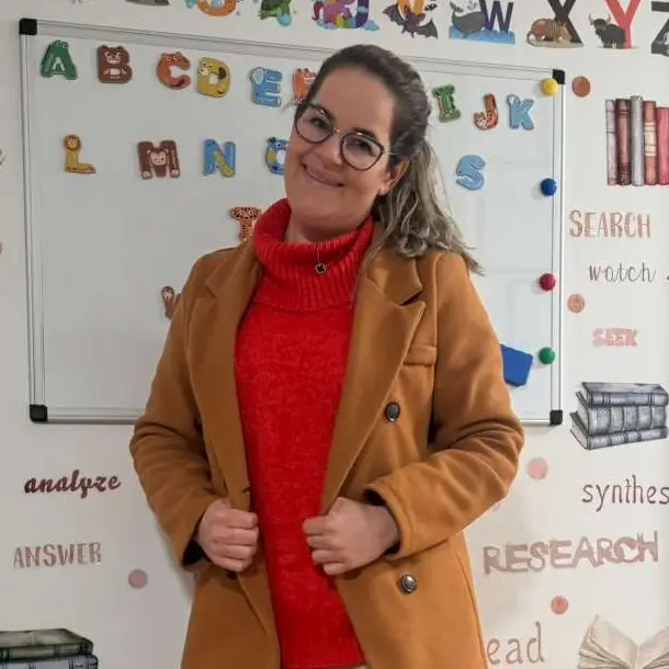 A Brazilian woman in a brown coat and red sweater standing in a daycare classroom.